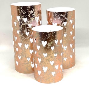May include: Three rose gold candle holders with a distressed finish and white heart cutouts. The candle holders are different sizes and are arranged in a row.