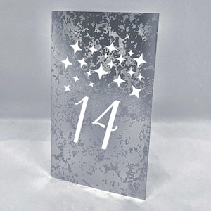 May include: A rectangular table number card featuring a mottled grey and silver textured background. The number 14 is printed in a large white serif font in the center, topped with a cluster of various sized white four-pointed star shapes.
