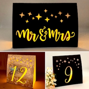 May include: A collection of three illuminated table signs. One black sign features the text Mr & Mrs with star cutouts. Another is a metallic gold sign with the number 12 and star cutouts. The third is a black sign with the number 9 and star cutouts.