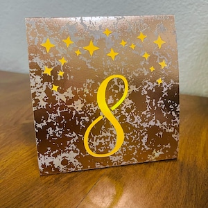 May include: A square table number card featuring a metallic copper and silver mottled background. The number 8 is printed in a large, bold yellow font in the center, surrounded by a cluster of small, yellow four-pointed star shapes at the top.