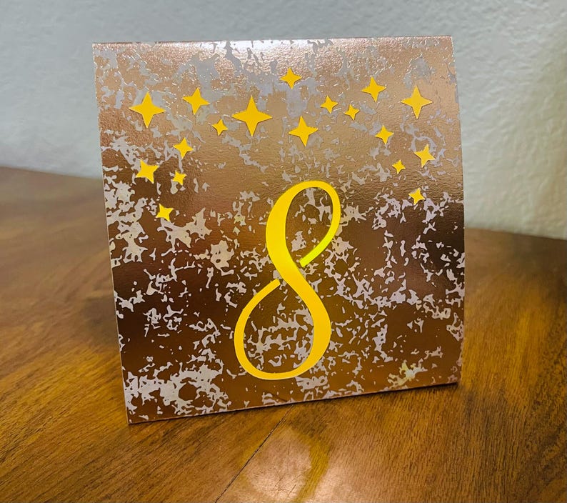 May include: A square table number card featuring a metallic copper and silver mottled background. The number 8 is printed in a large, bold yellow font in the center, surrounded by a cluster of small, yellow four-pointed star shapes at the top.