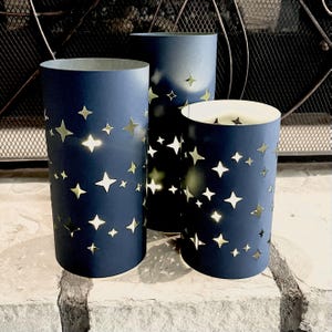 May include: Three cylindrical luminaries in dark navy blue, each featuring die-cut star shapes of varying sizes. The cutouts allow light to shine through from the interior, creating a glowing effect against the dark exterior surface of the cylinders.