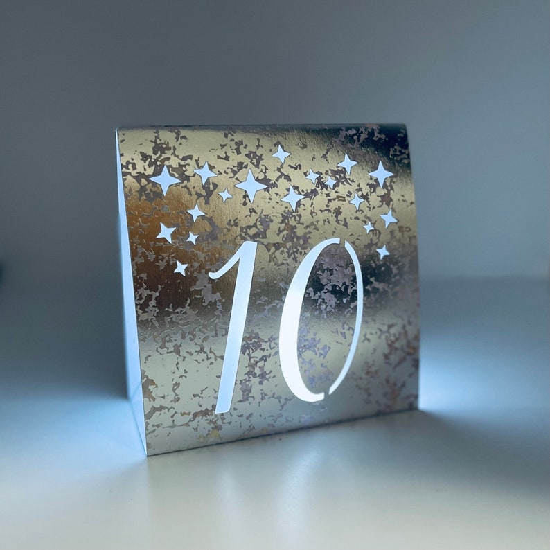 Star Design Night Table Number LUMINARIES are made with silver foil mercury glass style card stock and sheer vellum paper. The number and design is cut out for use with LED tea lights can shine through (not included.) The glowing numbers make it easy for guests to find their table.
