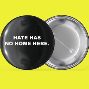 May include: A black button with the text "Hate has no home here." in white lettering. The back of the button is silver.