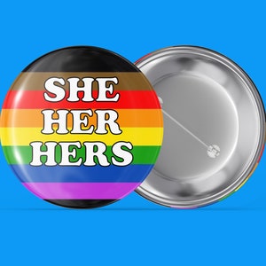 May include: A round button with a rainbow pride flag design and the text "SHE HER HERS" in white letters.
