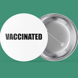 Könnte beinhalten: Zwei weiße runde Ansteckbuttons. Auf einem Button steht das Wort "VACCINATED" in schwarzen, fetten Buchstaben. Der andere Button zeigt die Rückseite mit einer silbernen Nadel. Der Hintergrund ist einfarbig teal.