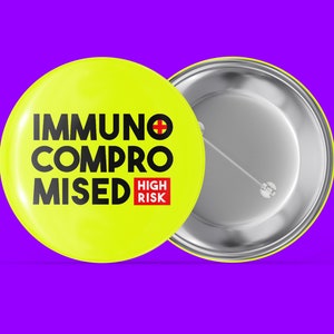 Könnte beinhalten: Gelber Knopf mit dem Text "Immunsupprimiert Hohes Risiko" in schwarzen Buchstaben mit einem roten Kreuzsymbol. Die Rückseite des Knopfes ist silber.
