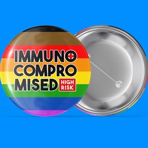 Könnte beinhalten: Ein regenbogenfarbener Knopf mit dem Text "Immunsupprimiert Hohes Risiko" in schwarzen Buchstaben. Der Knopf hat eine silberne Rückseite.