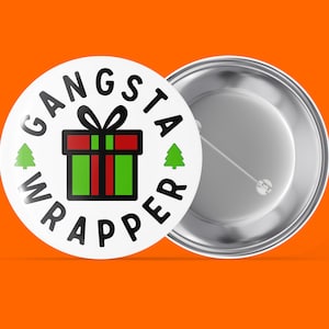 May include: A white button with a black and red gift box, green Christmas trees, and the text "Gangsta Wrapper" in black.