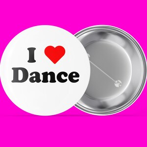 May include: A white button with a red heart and the text "I Love Dance" in black. The button is attached to a silver pin back.