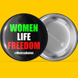 Op de afbeelding: Een zwarte knop met een witte pin aan de achterkant. De voorkant van de knop heeft de woorden "Women Life Freedom" in groene, witte en rode tekst. De tekst "#MahsaAmini" is in zwart onder de woorden gedrukt.