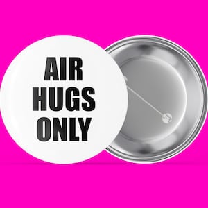 May include: White button with black text that reads "AIR HUGS ONLY". The back of the button is silver.