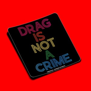 May include: Black sticker with the text "DRAG IS NOT A CRIME" in rainbow colors. The text is in a bold, stylized font. The sticker has a white border and the text "#MakeItLouder - EXPRESS YOURSELF" in smaller white text at the bottom.