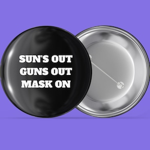 Könnte beinhalten: Ein schwarzer Knopf mit einer weißen Aufschrift, die "SUN'S OUT GUNS OUT MASK ON" lautet. Der Knopf befindet sich auf einem violetten Hintergrund.