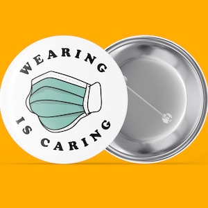 Könnte beinhalten: Ein weißer Knopf mit einer Schwarzweißillustration einer Gesichtsmaske und dem Text "Wearing is Caring". Die Rückseite des Knopfes ist silber.