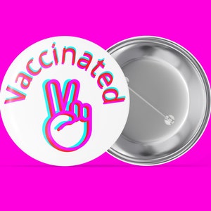 Peut inclure: Un badge blanc avec le mot "Vaccinated" en lettrage 3D rouge et bleu, et un signe de paix dans les mêmes couleurs. Le badge est posé sur un fond rose vif.