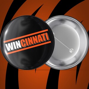 May include: A black button with an orange stripe and the text "WIN CINNATI" in white letters. The back of the button is silver.