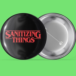 May include: A black button with a red and white design that says "SANITIZING THINGS" in a font similar to the Stranger Things logo.