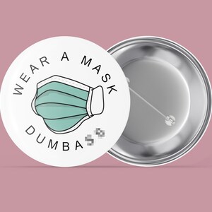 Könnte beinhalten: Weißer Knopf mit einer Schwarzweißillustration einer Gesichtsmaske und dem Text "WEAR A MASK DUMBA". Die Rückseite des Knopfes ist silber.
