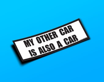May include: A rectangular bumper sticker with a white background and a thick black border. The text printed in bold black capital letters reads: MY OTHER CAR IS ALSO A CAR. The sticker is oriented at a slight angle against a bright blue background.