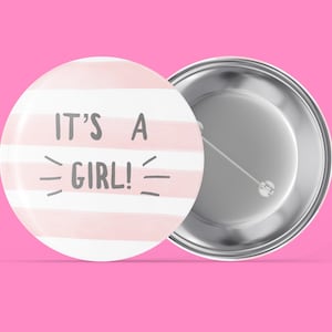 Può includere: Un bottone a strisce rosa e bianche con il testo "IT'S A GIRL!" in lettere grigie. Il retro del bottone è argentato.