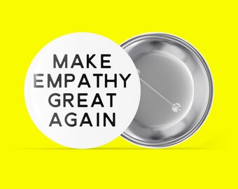 Puede incluir: Botón blanco con el texto negro "MAKE EMPATHY GREAT AGAIN". El botón tiene un alfiler plateado en la parte posterior y está sobre un fondo amarillo brillante. El botón es redondo.
