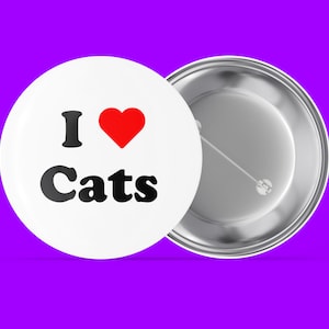 Puede incluir: Un botón blanco con un corazón rojo y el texto "I love Cats" en letras negras. El botón está unido a una parte posterior de pasador de plata.