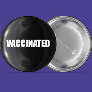 Könnte beinhalten: Ein schwarzer Knopf mit silberner Rückseite trägt die Aufschrift "VACCINATED" in weißer Schrift.