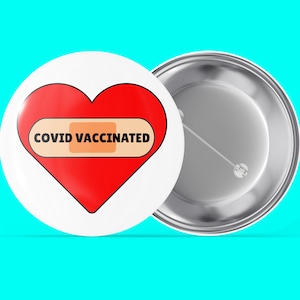 Könnte beinhalten: Ein weißer Knopf mit einem roten Herzen und einem Pflaster, auf dem "COVID VACCINATED" steht. Der Knopf ist an einer silbernen Rückseite befestigt.