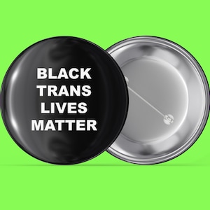 May include: Black button with the text "BLACK TRANS LIVES MATTER" in white letters. The back of the button is silver.