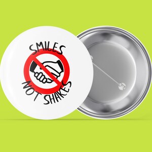 May include: A white button with a red circle and a diagonal line through a black handshake graphic. The text "SMILES NOT SHAKES" is written around the circle.