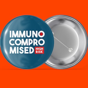 Könnte beinhalten: Ein blauer Knopf mit dem Text "IMMUNOCOMPROMISED HIGH RISK" in weißen Buchstaben mit einem roten Pluszeichen und den Worten "HIGH RISK" in rot. Die Rückseite des Knopfes ist silber.