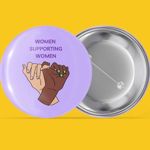 Puede incluir: Botón morado con el texto "Women Supporting Women" y un gráfico de dos manos que se sostienen. La parte posterior del botón es plateada.
