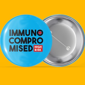 Könnte beinhalten: Ein blauer Knopf mit dem Text "IMMUNO COMPROMISED HIGH RISK" in Schwarz und Rot. Der Knopf hat eine silberne Rückseite mit einer Stecknadel.