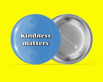 Puede incluir: Un botón azul con el texto "kindness matters" en letras blancas. El botón se muestra desde el frente y la parte posterior.