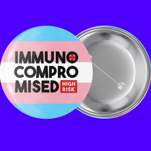 Könnte beinhalten: Ein runder Knopf mit einem Transgender-Pride-Flaggen-Design. Der Knopf trägt den Text "Immunsupprimiert Hohes Risiko" in schwarzer Schrift.