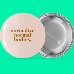 Può includere: Un bottone rosa con il testo "normalize normal bodies." in lettere marroni. Il retro del bottone è argentato.