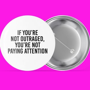 May include: A white button with a black text that reads "If you're not outraged, you're not paying attention". The button is on a pink background.
