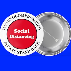 May include: White button with black text that reads "Immunocompromised Please Stand Back" with a red circle in the centre that says "Social Distancing".