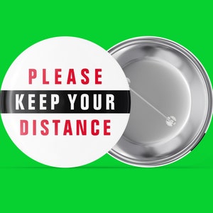 Könnte beinhalten: Ein weißer Knopf mit einem schwarzen Streifen und dem Text "PLEASE KEEP YOUR DISTANCE" in roten Buchstaben. Die Rückseite des Knopfes ist silber.
