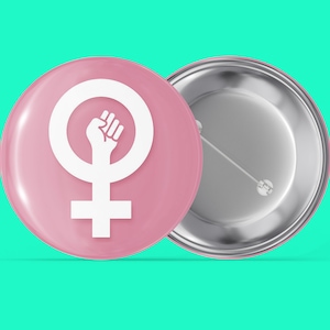 Puede incluir: Un botón rosa con un símbolo femenino blanco y un puño levantado dentro del círculo. La parte posterior del botón es plateada.