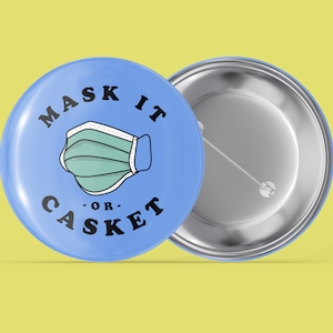 Könnte beinhalten: Ein blauer Knopf mit einer weißen Gesichtsmaske und dem Text "Mask It - Or - Casket" in Schwarz.