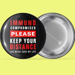 Könnte beinhalten: Schwarzer Knopf mit weißem Text, der "Immunkompromittiert Bitte Abstand halten Sie könnten mir das Leben retten" in Rot lautet. Der Knopf hat eine silberne Rückseite.