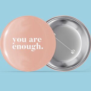 May include: A pink button with the text "you are enough." in white lettering. The back of the button is silver.