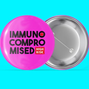 Könnte beinhalten: Zwei runde, pinkfarbene Ansteckbuttons. Einer zeigt den Text "IMMUNO COMPROMISED" mit einem roten Kreuzsymbol und "HIGH RISK" in einem roten Feld. Der andere Button zeigt die silberne Rückseite.