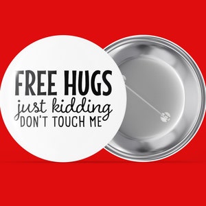 Könnte beinhalten: Weißer Knopf mit schwarzem Text, der "FREE HUGS just kidding DON'T TOUCH ME" lautet. Die Rückseite des Knopfes ist silber.