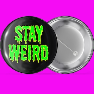 Puede incluir: Un botón negro con un texto verde que gotea que dice "STAY WEIRD". La parte posterior del botón es plateada.