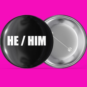 May include: A black button with a silver back. The button has the text "HE / HIM" in white letters.