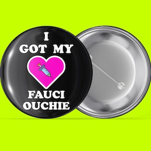 Könnte beinhalten: Ein schwarzer Knopf mit einem weißen Herzen, das eine Nadel enthält, und dem Text "I GOT MY FAUCI OUCHIE" in weiß.
