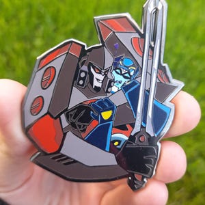 May include: A metal pin depicting two cartoon characters, one grey and red, the other blue and silver, in an embrace. The grey character holds a sword. The pin is held against a green background.